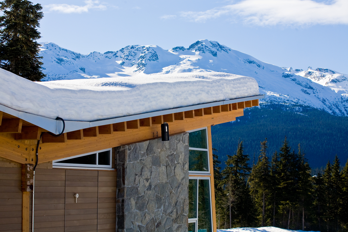 Outdoor sunny snowy view showing corner of hybrid wood building with angled glue-laminated timber (Glulam) beams supporting wooden roof structure, snow on top, and mountains in the distance
