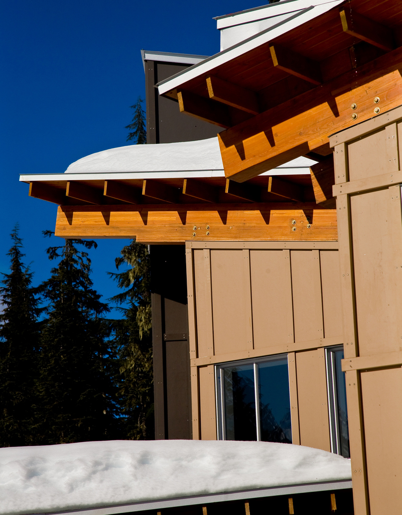 Outdoor sunny snowy view showing hybrid wood building with glue-laminated timber (Glulam) beams supporting wooden roof structure, snow on top, and trees in the distance -- all as an example of wood