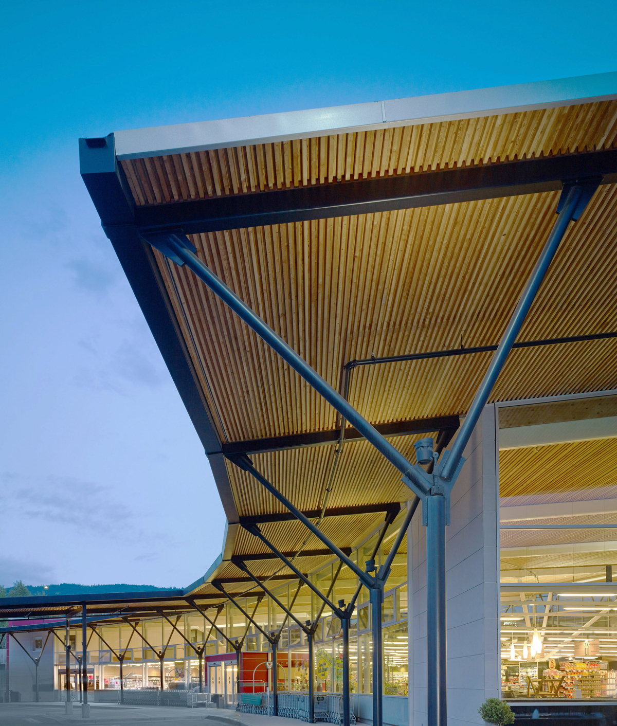 Exterior early evening view of Askews Supermarket front showing covered hybrid walkway of nail-laminated timber (NLT) and steel running along two storey low rise glass fronted store