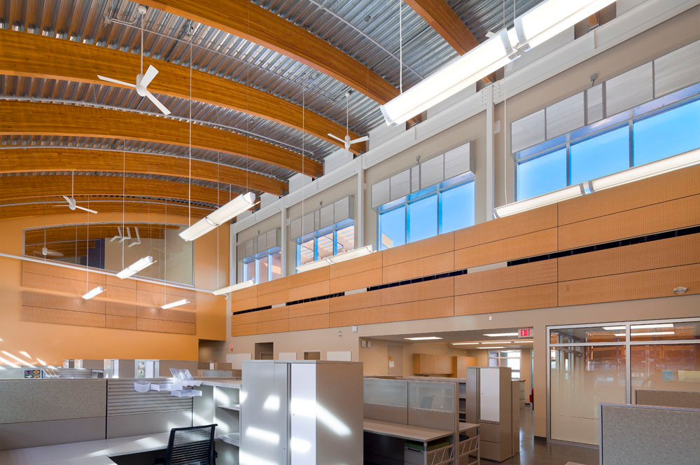 Daytime interior view of BC Hydro Maintenance and Operations Facility in Maple Ridge, BC showing office and work space with wood paneling and trim protected by curved glue-laminated timber (glulam) timber roof beams topped with a metal clad exterior roof surface visible above