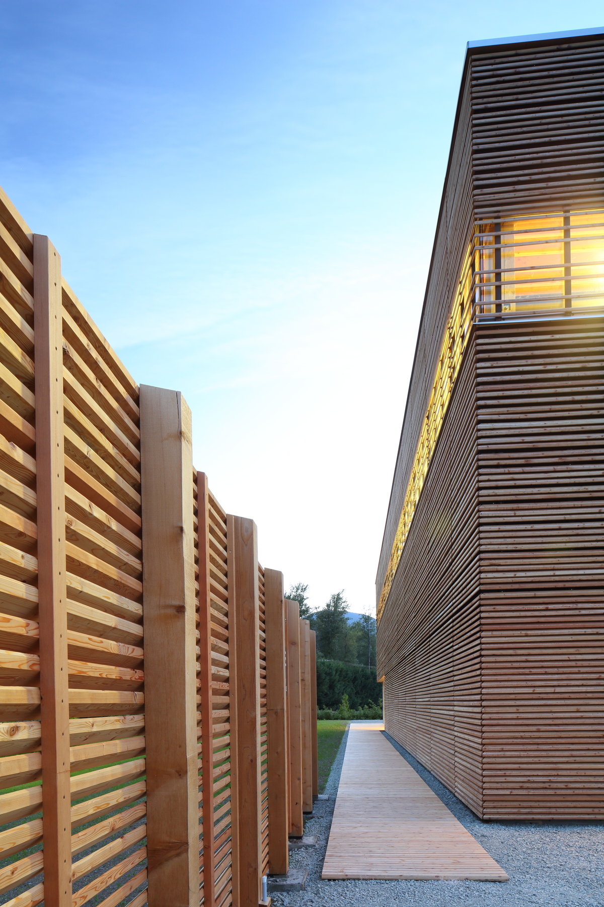 Exterior afternoon view of completed BC Passive House Factory, a low rise passive house structure built with light frame and mass timber components, showing wrap around exterior wood slats, wooden walkway, and decorative wooden fencing