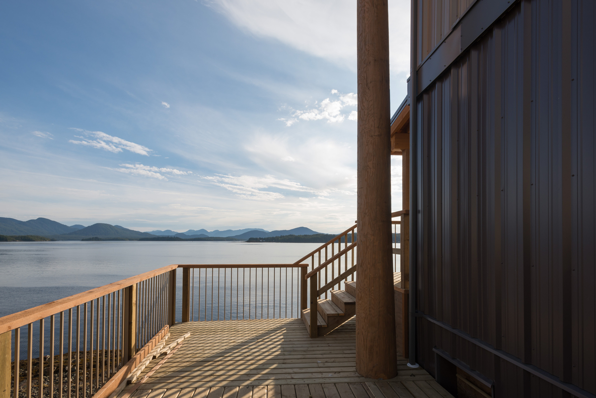 Exterior afternoon view of modular passive house / high performance Bella Bella Staff Housing, showing wood deck, wood railings, wooden stairs, vertical log pole, and metal exterior siding in foreground with Lake Agassiz and mountains in distance