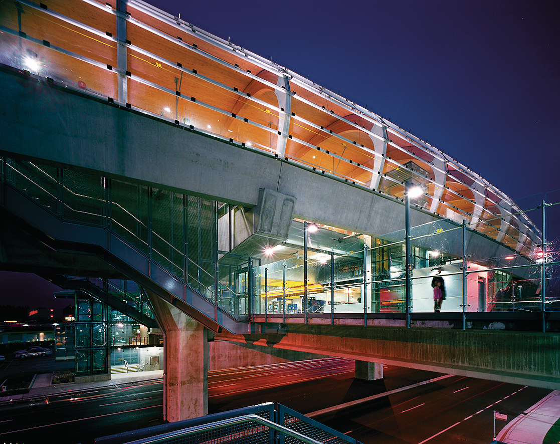 Curved glass exterior panels and large arching nail laminated timber (NLT) beams supporting a wooden ceiling are prominent features in this artistic night time view of the City of Burnaby's Brentwood Town Centre Transit station