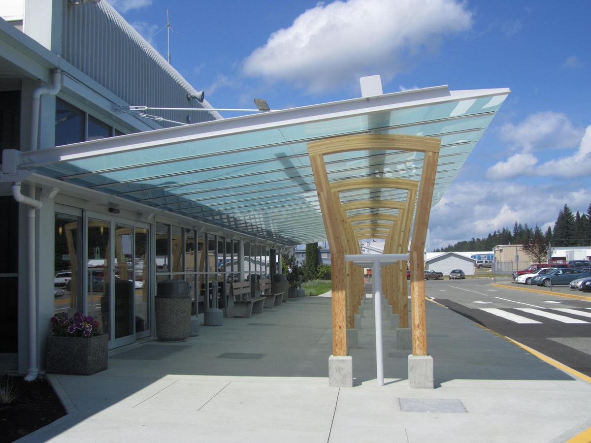 Exterior sunny daytime view of covered entrance to lowrise Campbell River Airport Expansion showing glulam 'arch' structures which support a large glass canopy that creates an outdoor entry room