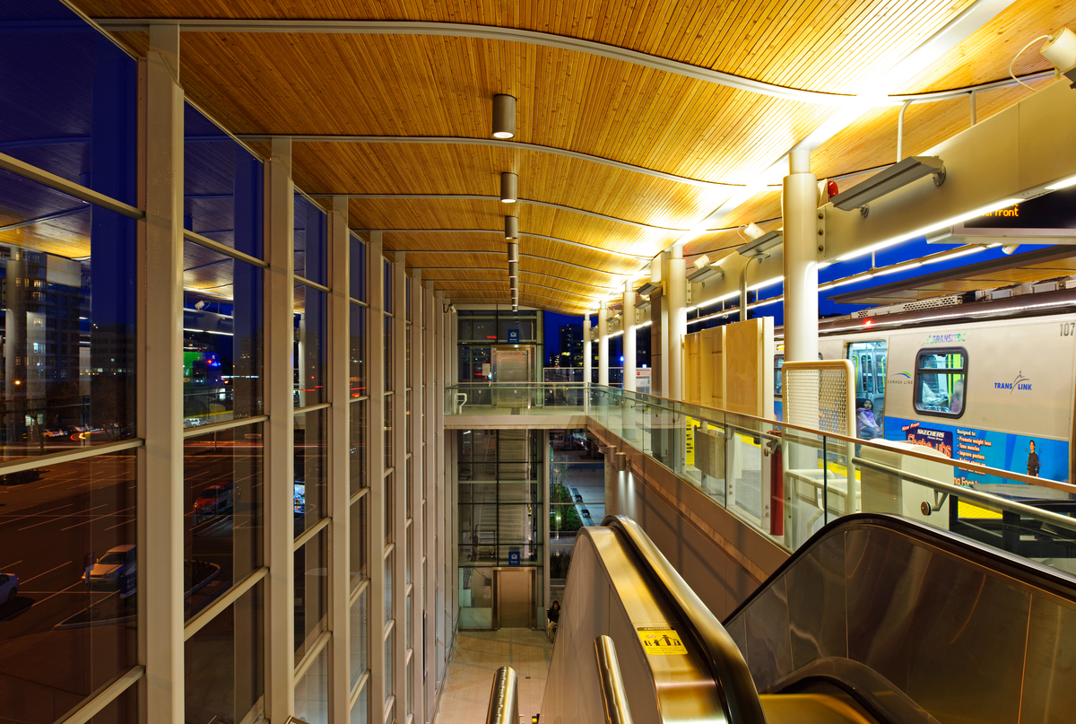 Lansdowne Canada Line Station in evening with wood detail ceiling, pot lights and skytrain in station. Escalator in foreground.