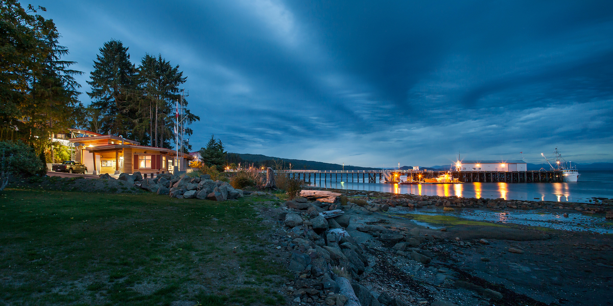 Exterior distant evening view of Canadian Coast Guard Search and Rescue Station showing shoreline, rocks, water, pier, and the station proper with exterior wood siding, post & beams, and wood trim