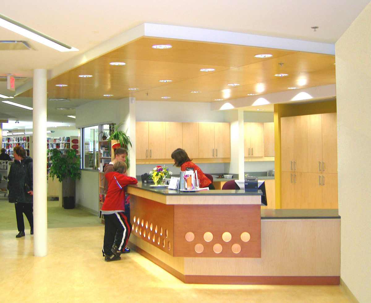 Interior daytime view of low rise Cranbrook Public Library, showing populated library, reception desk and abundant millwork, including reception desk, ceiling paneling, cabinetry, and decorative elements