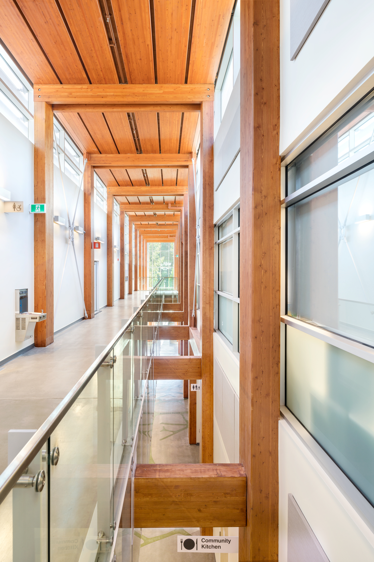 Interior view of Delbrook Community Recreation Centre multi storey main hallway from top floor, showing aesthetic wood planking overhead supported by glue-laminated timber (glulam) beams and columns