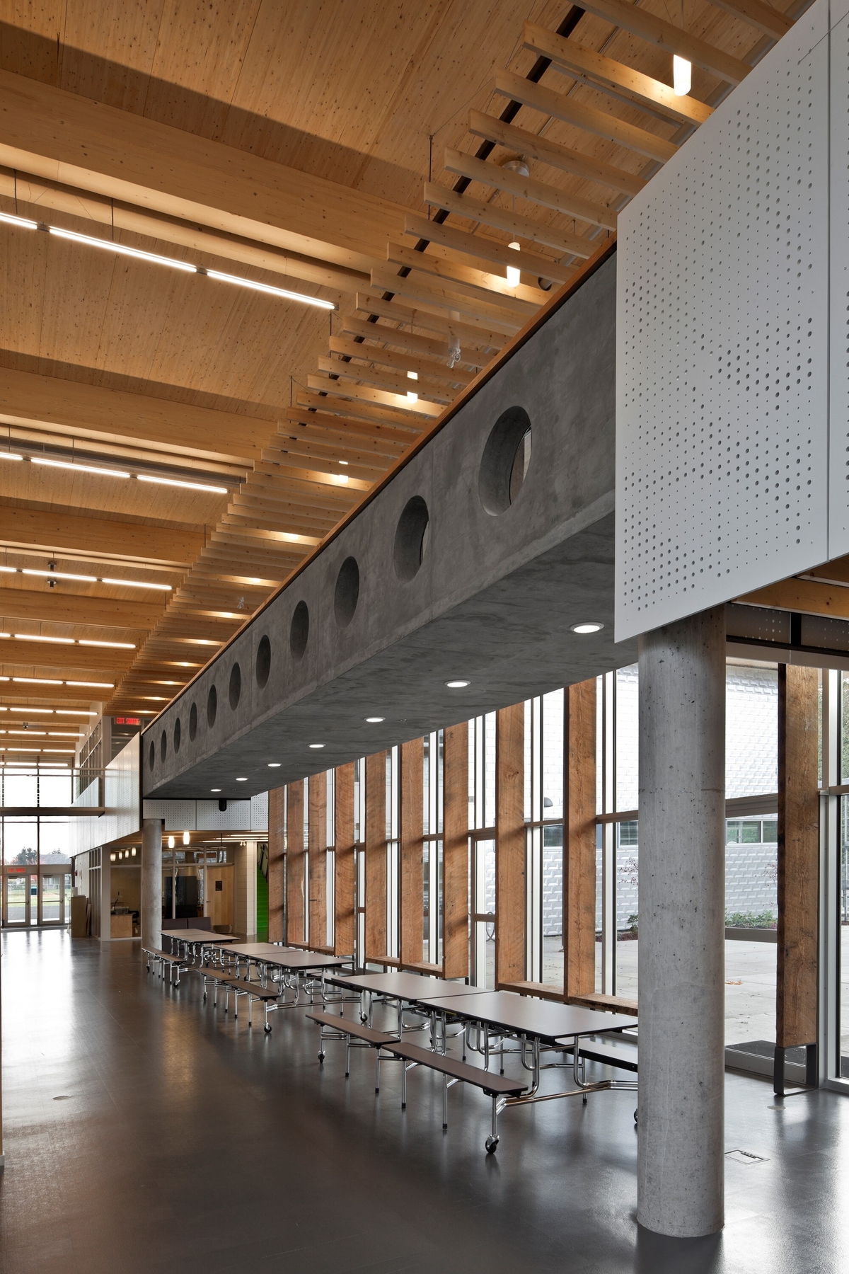 Interior daytime view of low rise École Au-cœur-de-l’île elementary school two storey main entrance hall and office, showing exposed Douglas-fir timbers, glue-laminated timber (glulam) roof beams, 2nd floor concrete walkway, and extensive wood accents