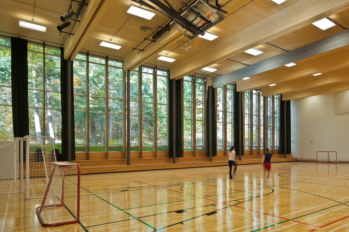 Sunny daytime interior view of École Mer-et-Montagne gymnasium showing entire exterior glass wall, exposed Douglas-fir ceiling beam timbers supporting CLT ceiling sections, all above a wooden multi-purpose floor occupied by 2 students