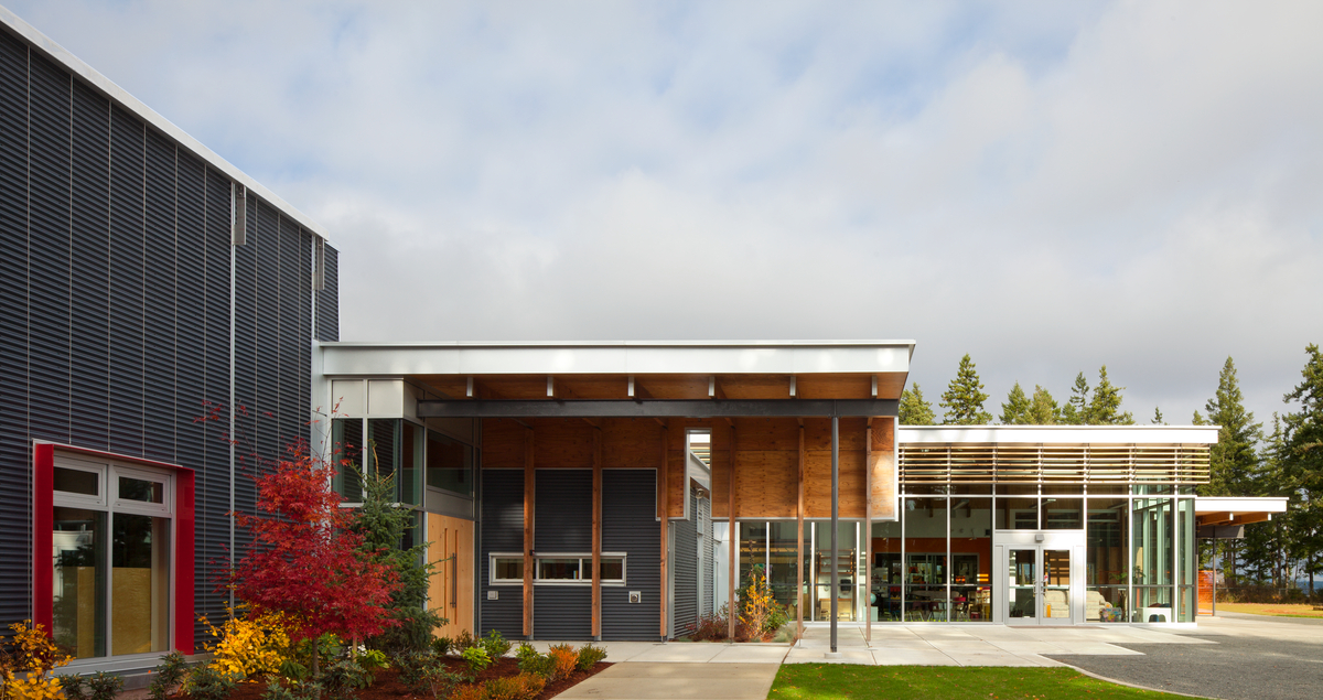 Exterior daytime view of low rise hybrid École Mer-et-Montagne elementary school, showing glass fronted library and large wood roof as part of covered entrance