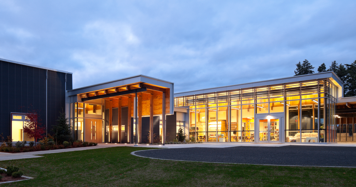 Exterior evening view of low rise hybrid École Mer-et-Montagne elementary school, showing glass fronted library and large wood roof as part of covered entrance
