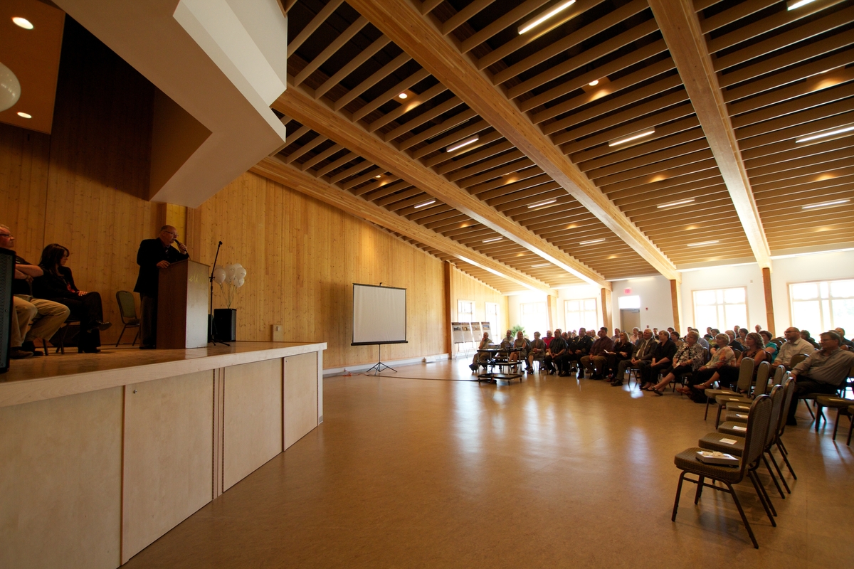 Interior daytime view of seated attendees and speaker at podium within Elkford Community Conference Centre main hall, the hall includes structural elements of glue-laminated timber (Glulam) and Laminated veneer lumber (LVL) beams, along with cross-laminated timber (CLT), providing the visible wall and ceiling/roof structure