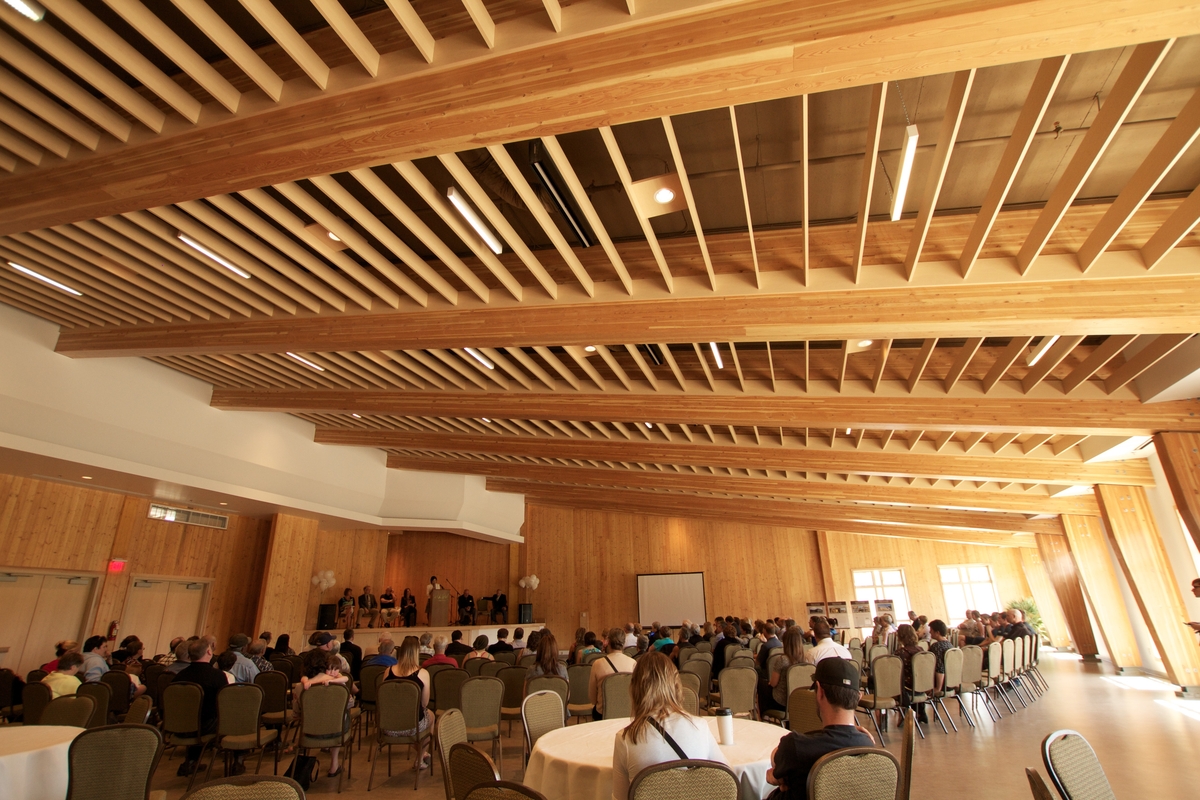 Interior daytime view of numerous seated persons attending conference inside Elkford Community Conference Centre with structural elements of glue-laminated timber (Glulam) and Laminated veneer lumber (LVL) beams, along with Cross-laminated timber (CLT), providing the wall and ceiling/roof structure