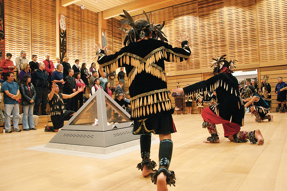 Interior view of The First Peoples House at the University of Victoria showing First nations people in traditional regalia dancing in a warmly lit room with extensive use of wood wall paneling, wood flooring, and mass timber, including Douglas-fir glue-laminated timber (glulam) beams