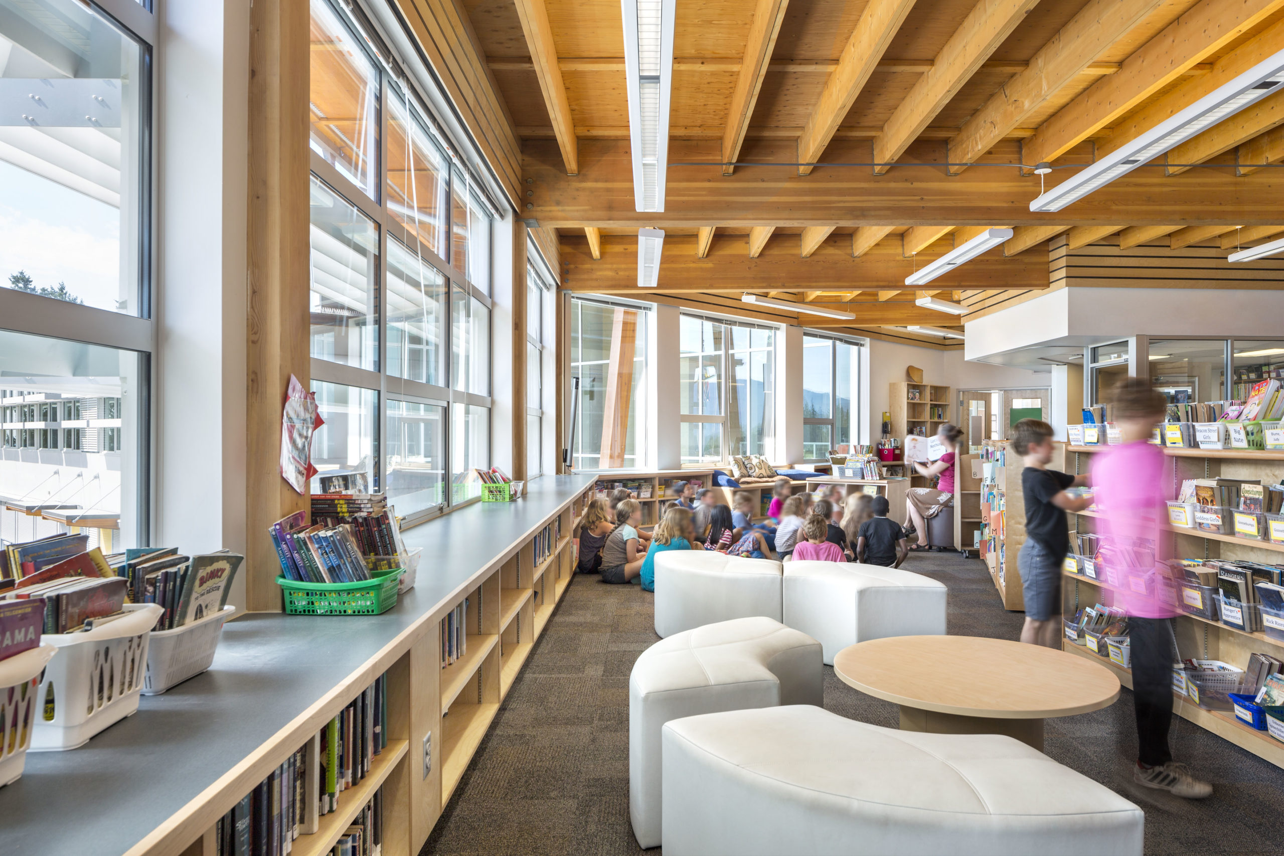 Interior view of elementary school library with exposed wood beams in ceiling, windows across most walls, and kids grouped on the floor listening to an instructor with a book.