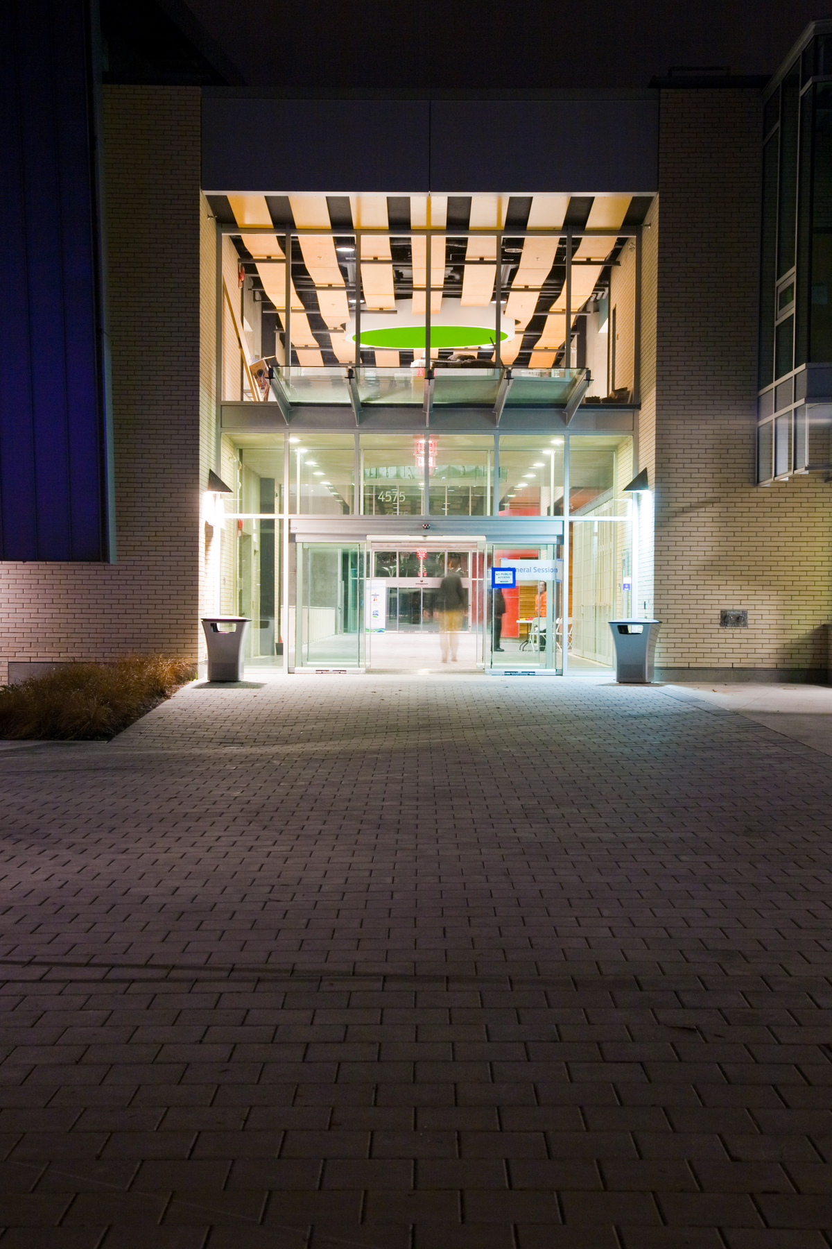 Exterior nighttime image of low rise Hillcrest Centre main entrance showing large glass windows, exterior glass awning, and interior decorate undulating ceiling 'waves' of wood