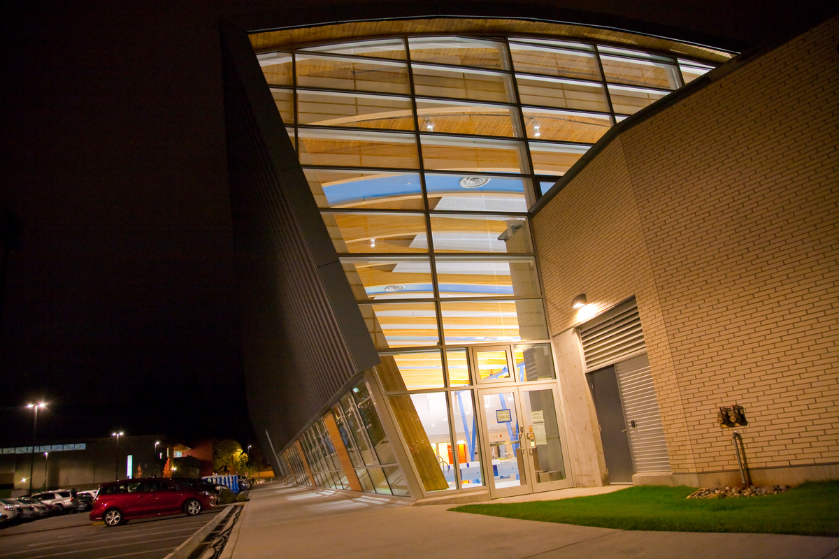 Exterior nighttime image of low rise Hillcrest Centre showing large glass expanses and ceiling of exposed wood decking and a series of gently curved glue-laminated timber (glulam) beams inside