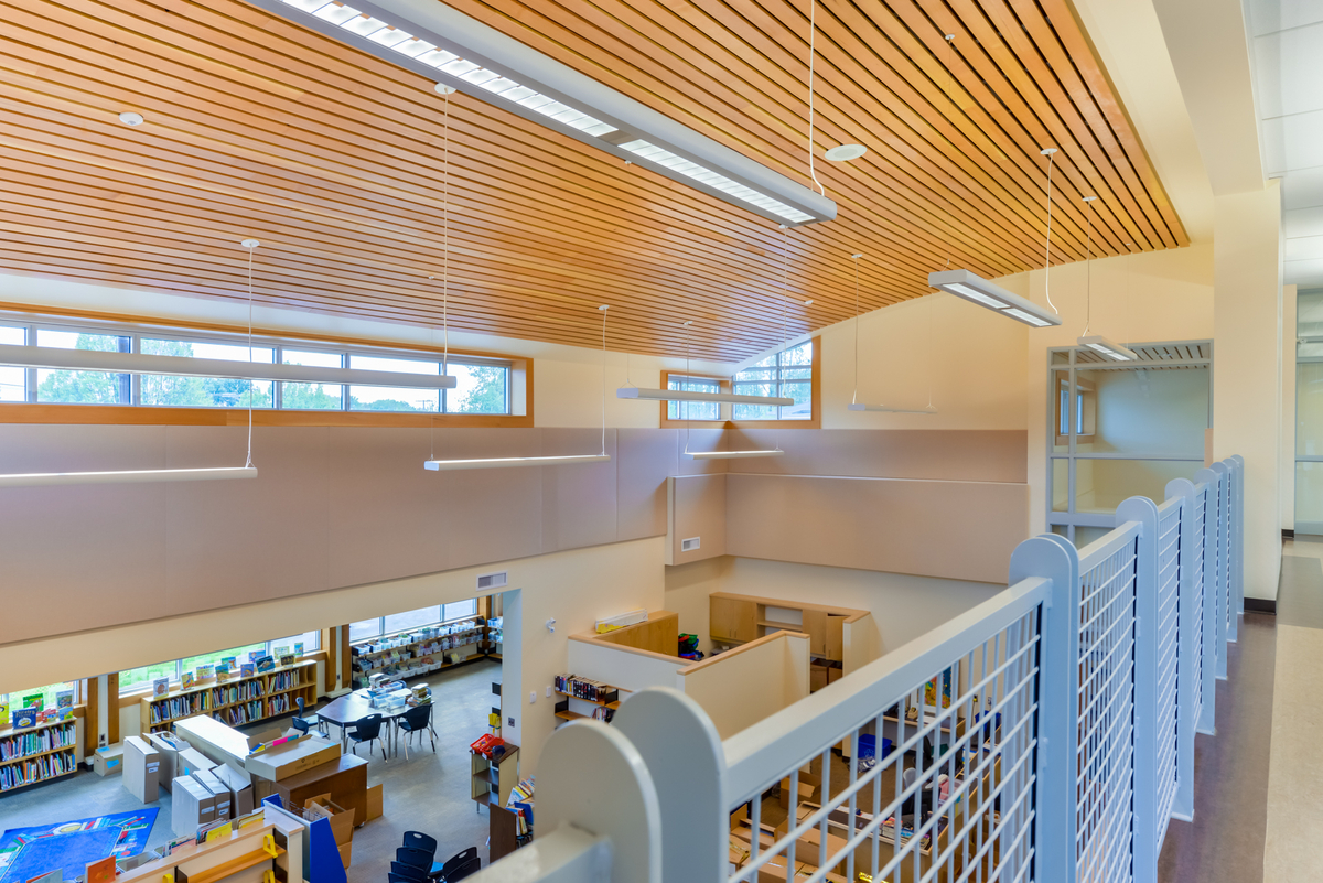 Indoor daytime view of low rise J.W. Sexsmith Elementary School library, from second floor balcony, showing the wood slat acoustic ceilings used in the library and other noise-sensitive area