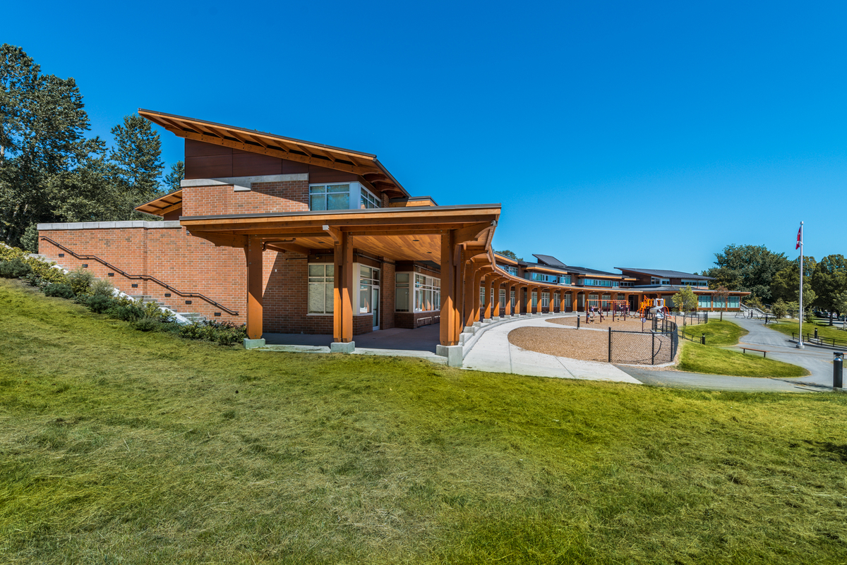 Exterior sunny daytime view of J.W. Sexmith Elementary School showing sweeping curved mass timber covered walkway along building front which features glue-laminated timber (glulam) beams, parallel strand lumber (PSL), and Solid-sawn heavy timber