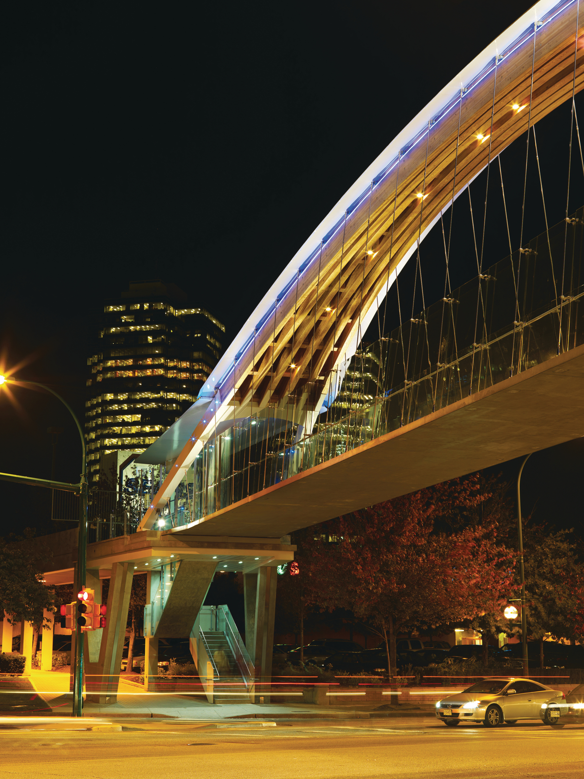 Exterior nightime view of Kingsway Pedestrian Bridge showing double-curved glue-laminated timber (glulam) arch with walkway suspended by steel cables below