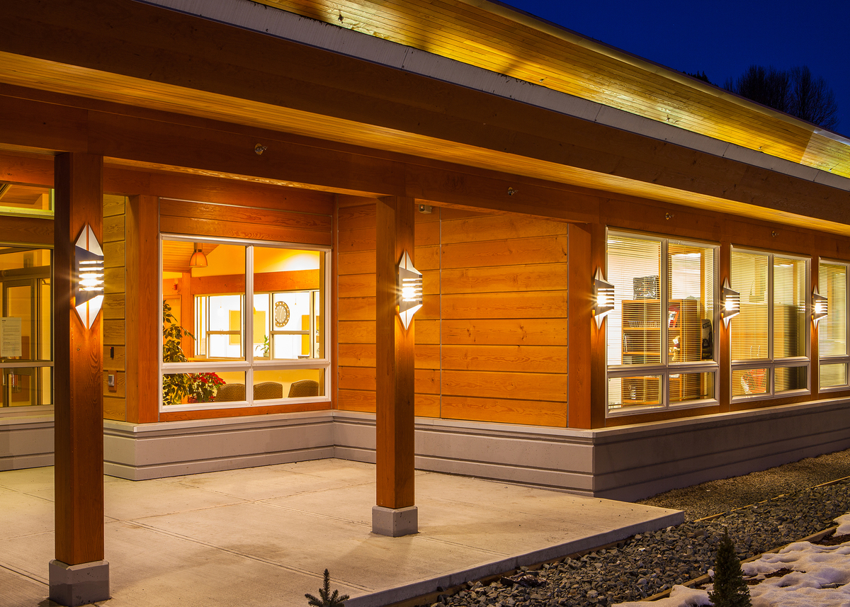 Exterior evening view of low rise Kitsumkalum Health Centre, showing extensive use of wood components, including paneling, plywood, siding, and solid-sawn heavy timber in both Douglas-fir and western red cedar