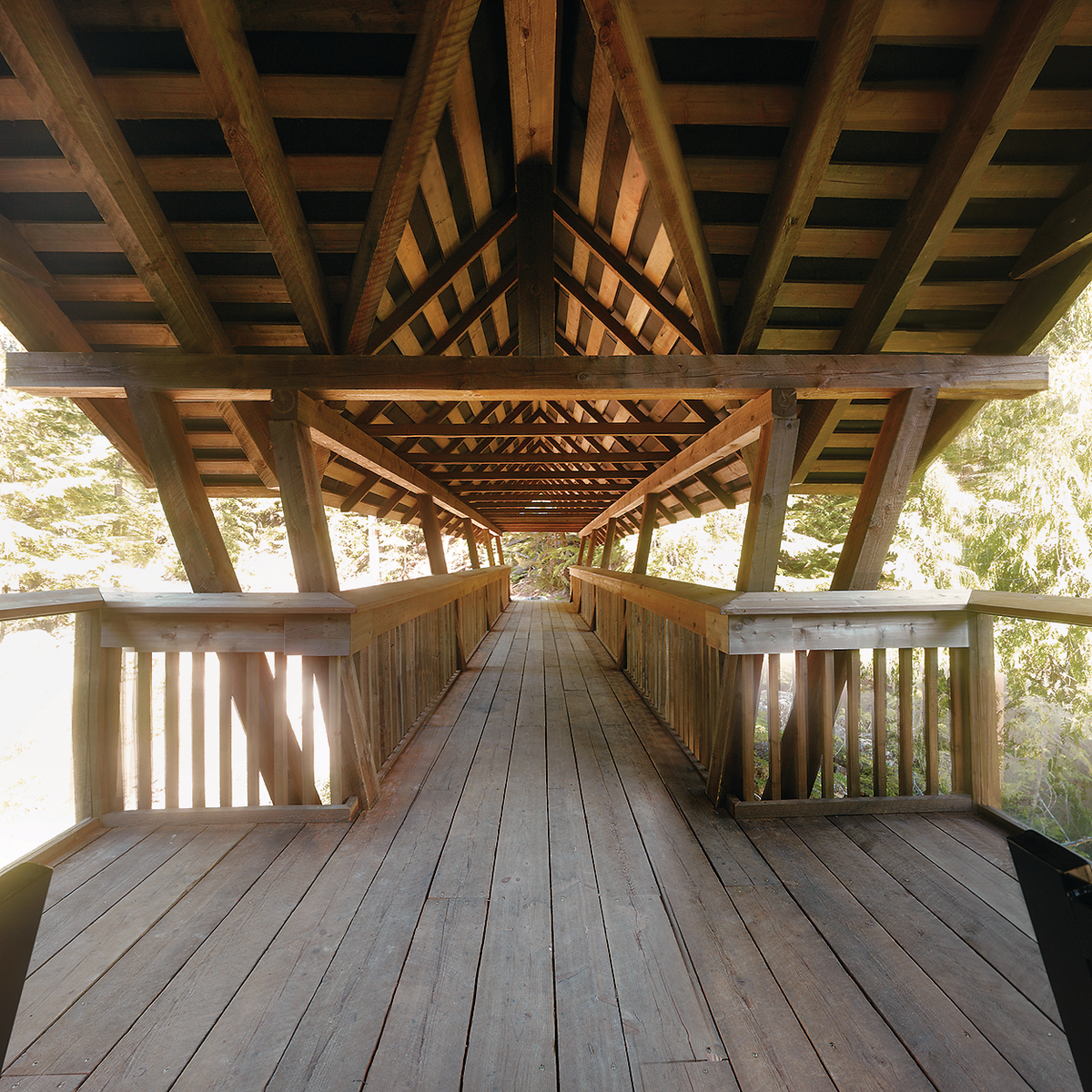 Daytime mid span view of Kuskanax River Footbridge showing wooden bannisters, railings, and roof structure constructed of glue-laminated timber (Glulam), Lumber, and solid-sawn heavy timber