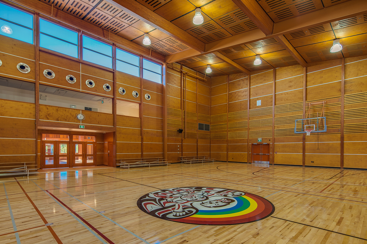 Sunny daytime interior view of Kwakiutl Wagalus School gymnasium showing extensive use wood, including western red cedar in posts, beams, and cladding, Douglas-fir for doors and windows, birch wood veneer finishes, and a maple hardwood sports floor