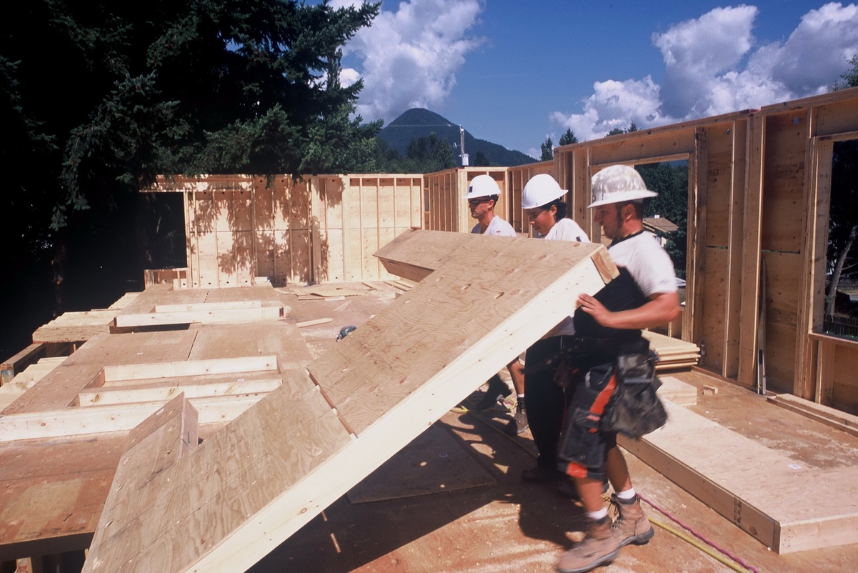 Sunny mid day image showing three workers tilting up prefabricated exterior light-frame wooden wall panel on construction site