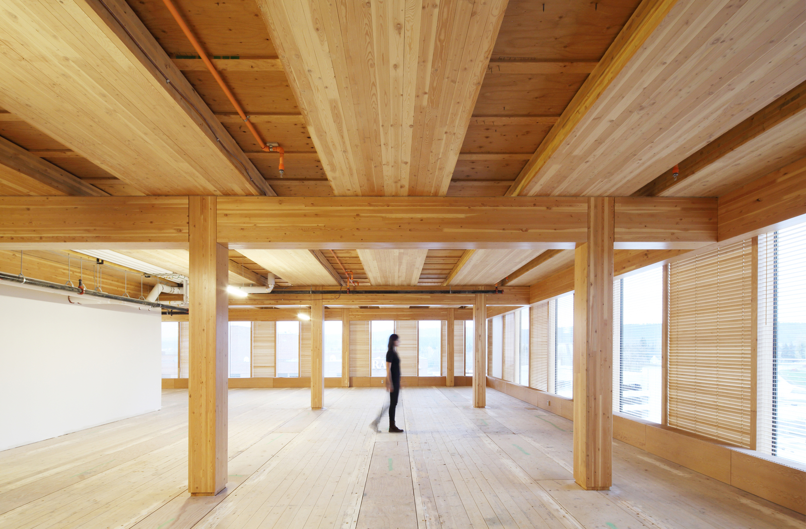 Interior daytime view of woman walking in multi storey mass timber structure showing extensive use of wood, including glue-laminated (glulam) timber beams and columns supporting plywood subfloor above and hardwood flooring below