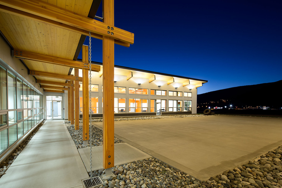 Exterior nighttime view of low rise Merritt Fire Zone Office and Provincial Wildfire Training Centre showing cross-laminated timber (CLT) covered entrance walkway stretching along glass exterior