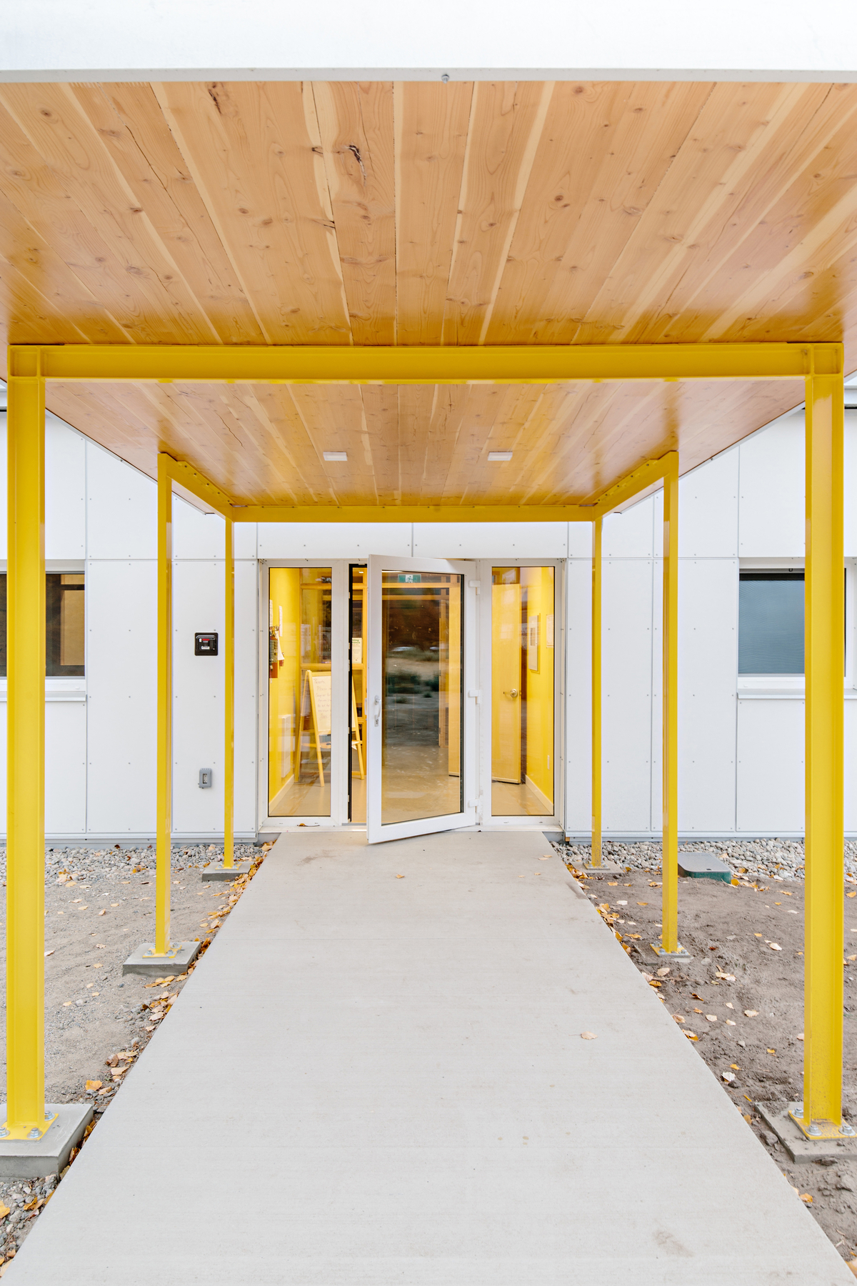 Exterior cloudy daytime view of low rise Okanagan College Child Care Centre entrance with yellow painted steel supports showing hybrid construction with light frame and mass timber construction, including wood covered entranceway