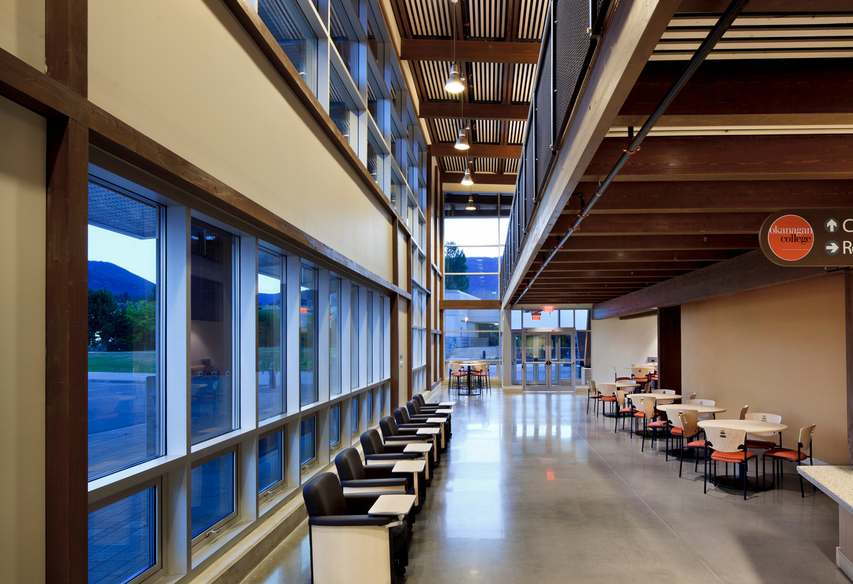 Interior evening view of low rise Okanagan College Jim Pattison Centre Of Excellence interior hallway showing glass exterior walls, glue-laminated timber (Glulam) supported balcony, and main floor seating area