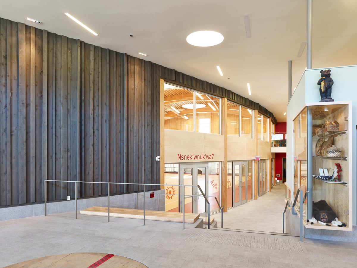 Interior daytime view of two-storey P’Egp’Ig’Lha Community Centre showing concrete and wood steps leading down to curved glass and natural wood siding of gymnasium entrance
