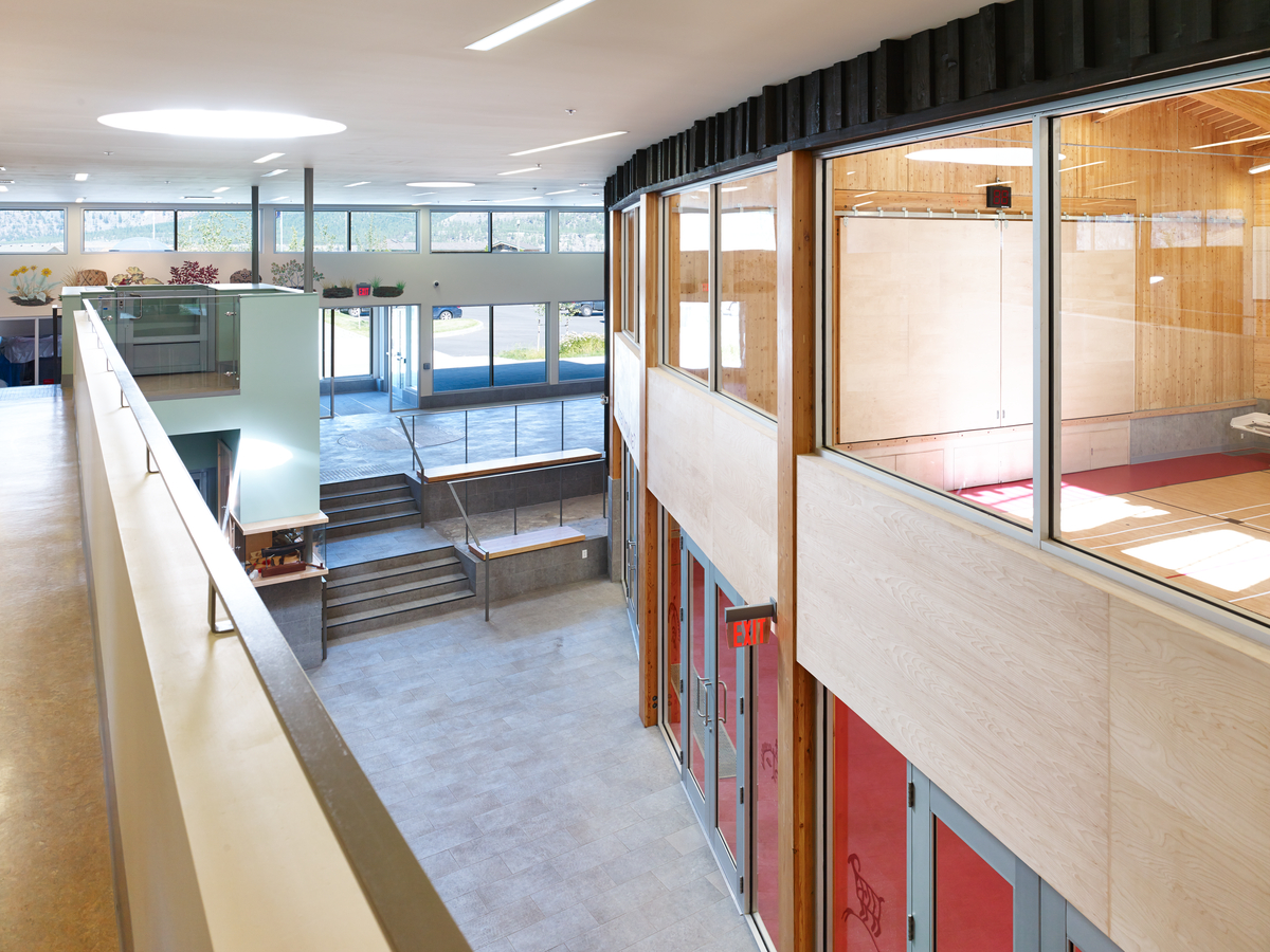 Interior daytime balcony view of two-storey P’Egp’Ig’Lha Community Centre showing concrete entrance steps leading to lobby with vertical glue-laminated timber (glulam) posts, shallow glulam arches, and cross-laminated timber (CLT) panels