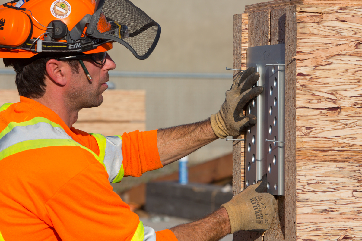 Exterior daytime close up view of worker in PPE using metal WIDC drilling guide on parallel strand lumber (PSL) beam