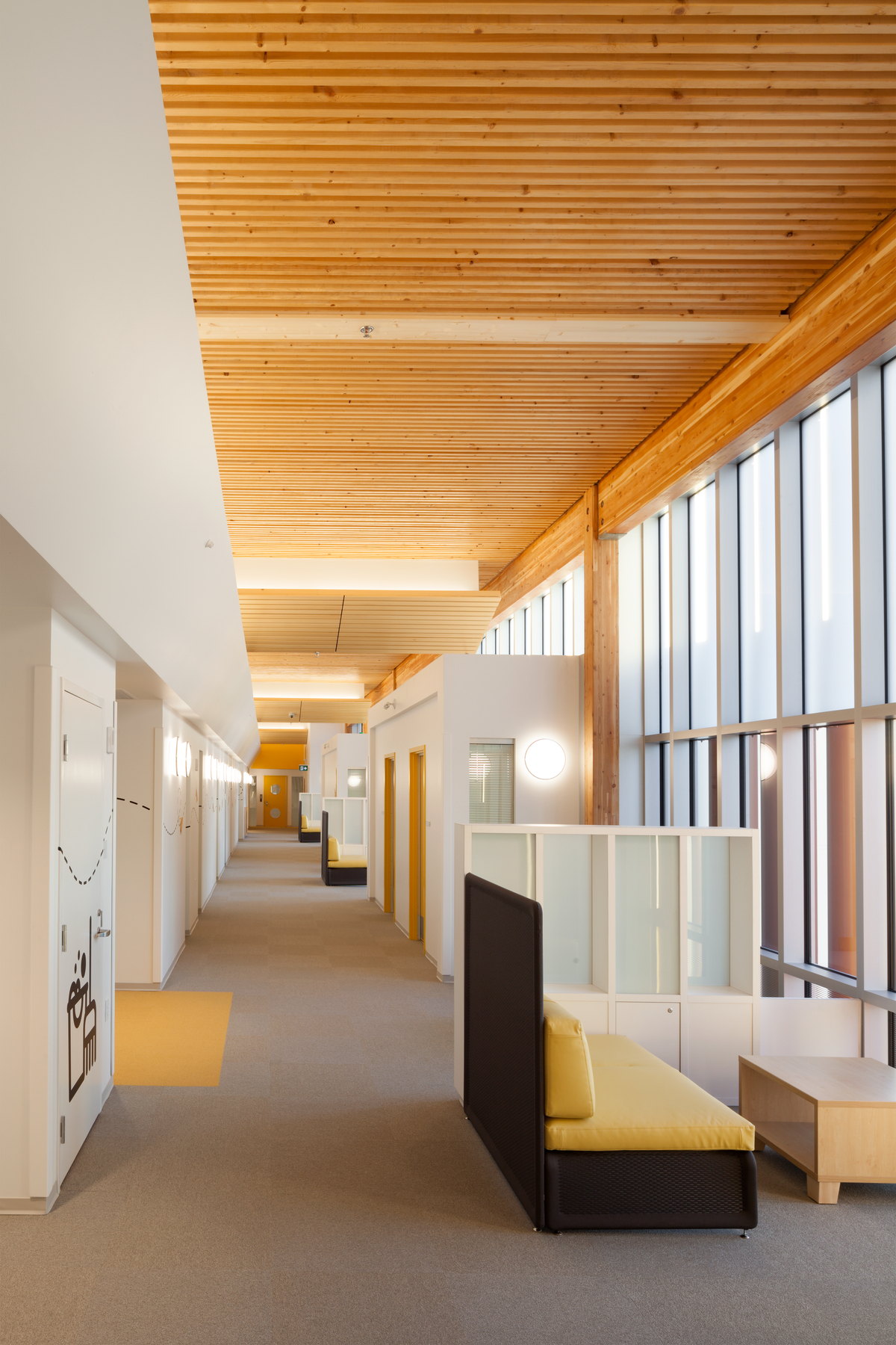 Bright sunny interior view of Pacific Autism Family Centre main hallway, showing exterior glass windows, and interior wood which includes glue-laminated timber (glulam) beams and columns, wood cabinets, wood trim, and dimensional lumber ceiling above