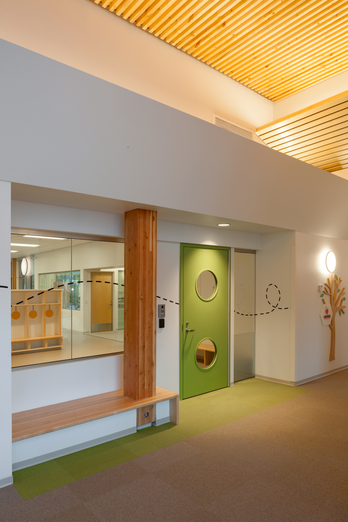 Bright sunny interior view of Pacific Autism Family Centre, showing colorful green interior door near glue-laminated timber (glulam) columns supporting dimensional lumber slat ceiling above