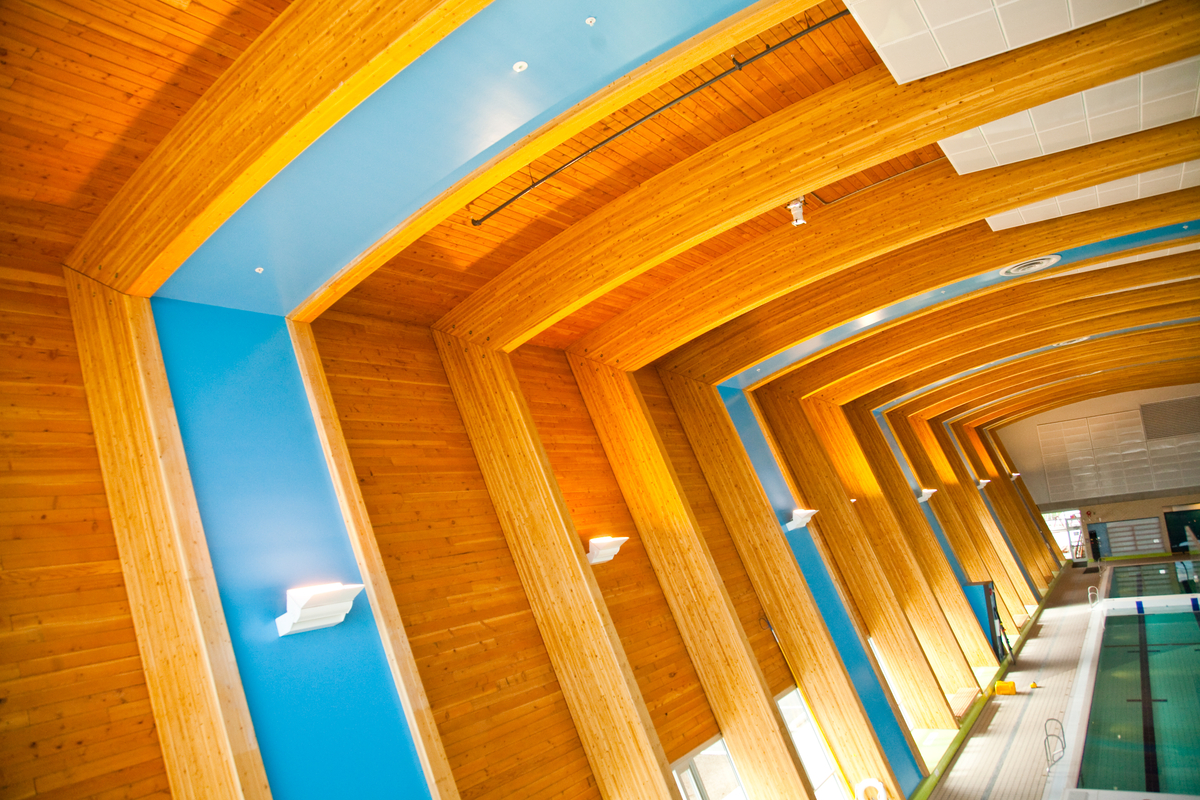 Interior daytime image of low rise Hillcrest Centre family aquatic main pool area showing ceiling of exposed wood decking and a series of gently curved glue-laminated timber (glulam) beams seamlessly connected to a series of glulam columns
