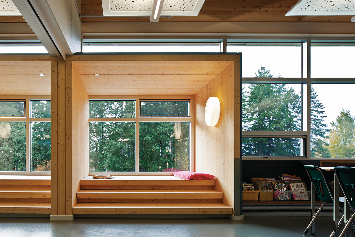 Interior daytime view of low rise École Au-cœur-de-l’île elementary school classroom, showing large glass window framed study pod with exposed Douglas-fir stairs, wall panels, and roofing