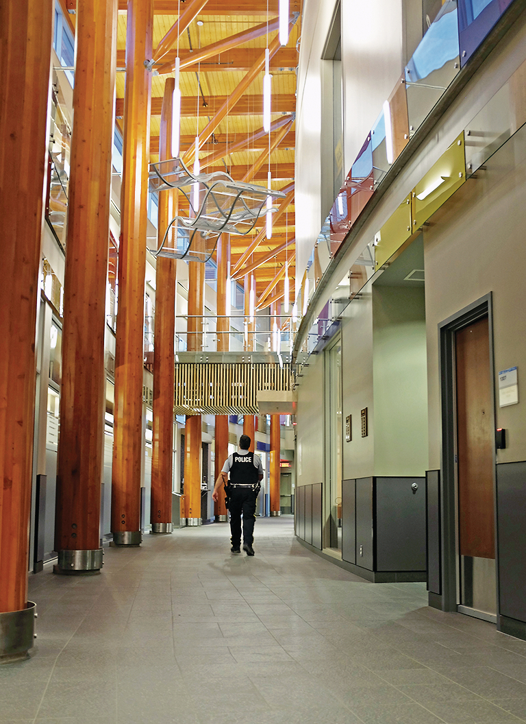 Interior daytime view of three-storey atrium of low rise Prince George RCMP Detachment showing post + beam, wood paneling, hybrid elements, and RCMP officer