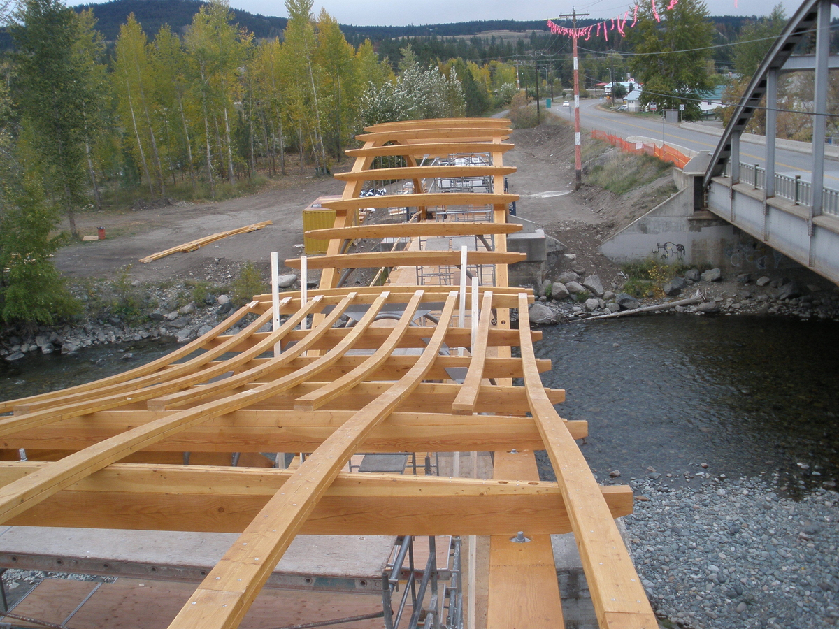 Outdoor overcast inline mid construction aerial view of Princeton Bridge of Dreams showing large glue-laminated timber (glulam) arches and undulating partially completed roof of wood and steel sheathing