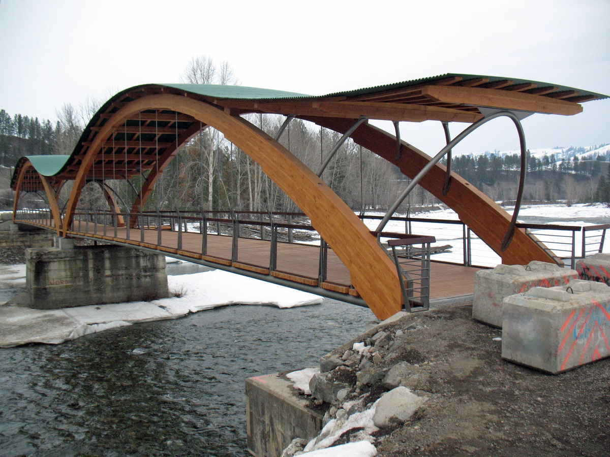 Outdoor overcast snowy full expanse profile view of Princeton Bridge of Dreams showing large glue-laminated timber (glulam) arches, wooden decking, and undulating roof of wood and steel sheathing