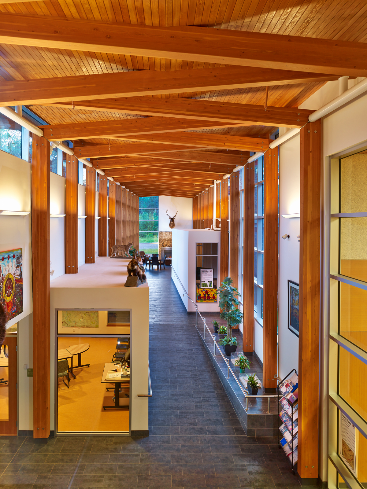 Interior daytime image of brightly lit Prophet River Multiplex main atrium showing downward tile walkway, glue-laminated timber (glulam) columns, crossed ceiling beams, and wood ceiling decking