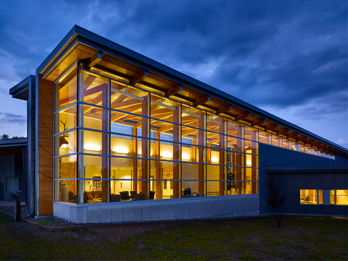 Exterior evening view looking into wide glass expanses of Prophet River Multiplex showing Douglas-fir glue-laminated timber (glulam) post-and-beam system which makes up the primary structure in this large linear lobby
