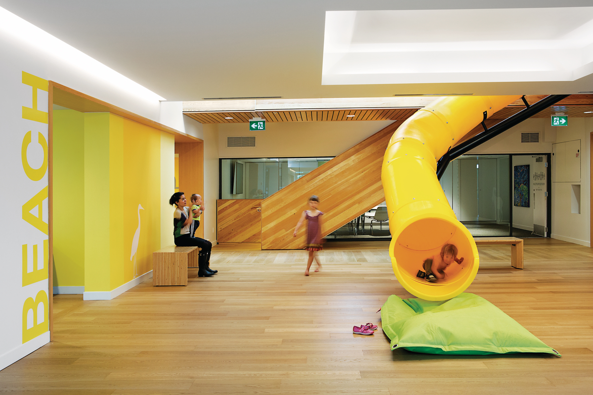 Interior daytime view of common space within the Yukon BC Ronald McDonald House showing children arriving into common space by way of a wooden staircase and an enclosed yellow tube slide