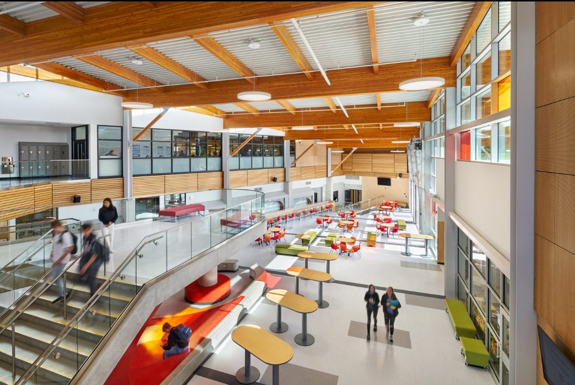 Interior daytime occupied view of multi storey École Salish Secondary School showing colorful main atrium with students, tables, and chairs below; and glue-laminated timber (glulam) beams above