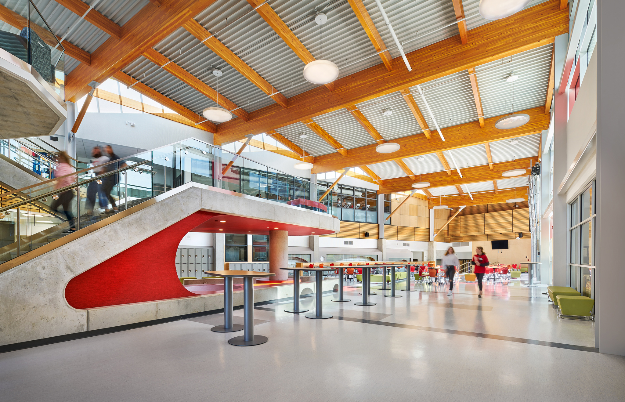 Interior daytime occupied view from ground floor of multi storey École Salish Secondary School showing colorful main atrium with students, tables, and chairs; and glue-laminated timber (glulam) beams above