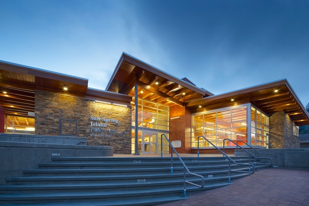 Exterior early evening view of low-rise Salt Spring Island Library showing exterior stairs leading up to glass, rock, and wood building with glue-laminated timber rafters extend beyond the north and south walls, visible beneath the cantilevered eaves