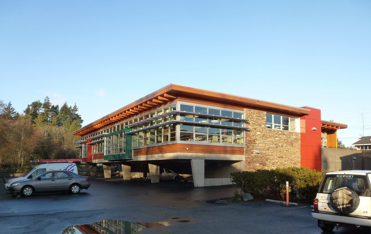 Exterior early evening view of low-rise Salt Spring Island Library showing wood, rock, glass, and concrete building exterior topped with glue-laminated timber rafters which extend beyond the north and south walls and are visible beneath the cantilevered eaves